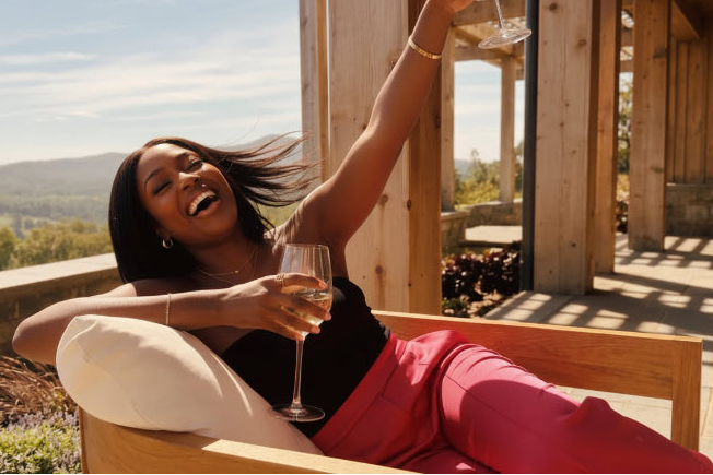 Woman in fushia satin flare pants sittingon a wooden bench outdoors, holding a glass of wine.slider_item_VBxtca
