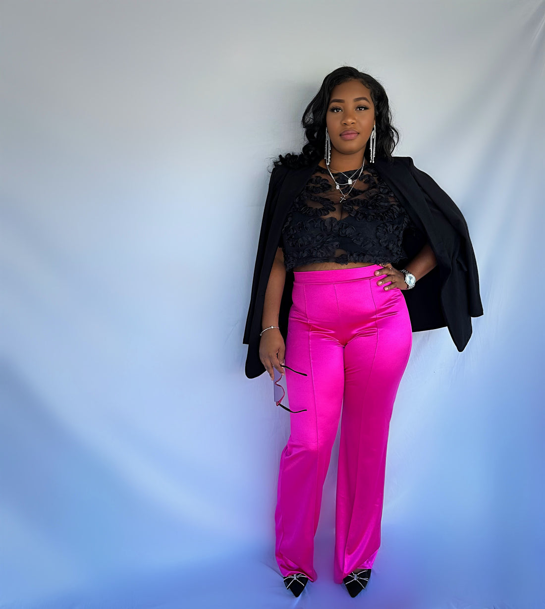 A model wearing bright pink high waisted pleated pants with a back zipper, paired with a black top and accessories.
